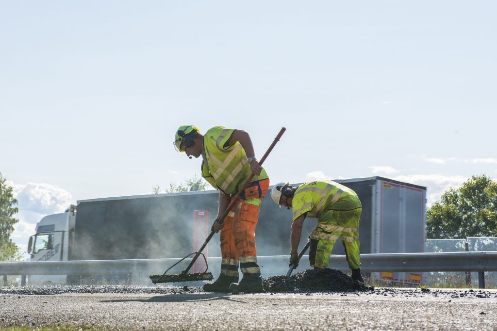 Två vägarbetare står och arbetar med asfaltering intill en väg där en lastbil kör förbi.