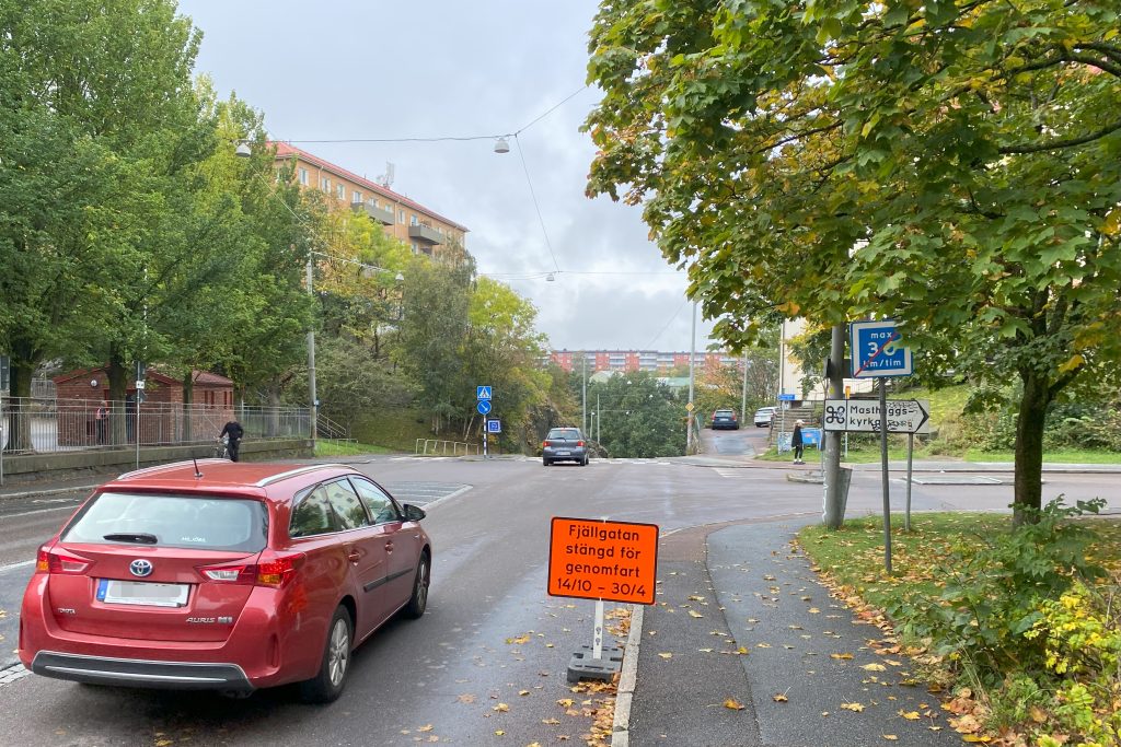 Två personbilar kör längs Fjällgatan i Masthugget. Fotgängare går med sin cykel på trottoar vänster i bild. En orange vägskylt med info om avstängningen syns vid sidan av vägen.