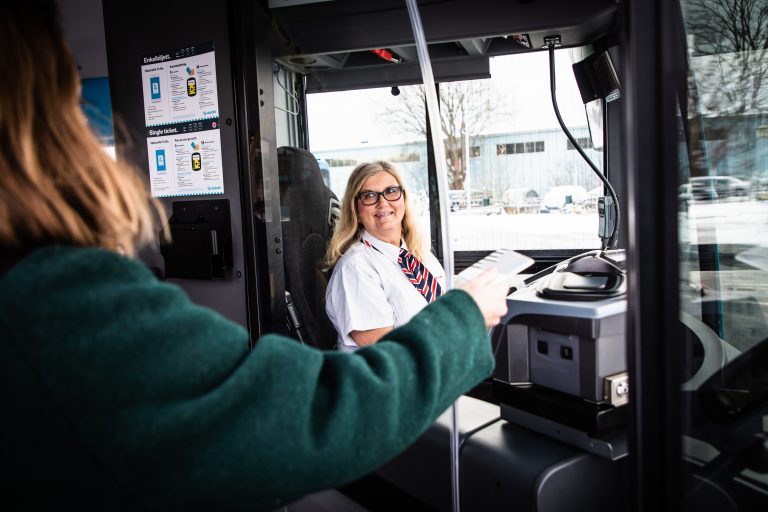 En kvinnlig busschaufför ler mot en passagerare som står med ryggen mot kameran.