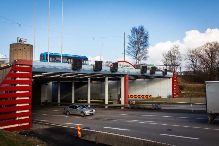 En personbil kör ut från Tingstadstunnelns mynning på fastlandssidan. En buss kör på vägen ovanför tunneln.
