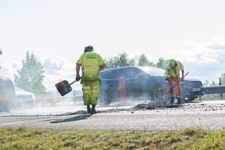 Beläggningsarbete det ryker om den heta asfalten där två personer står och arbetar bland passerande bilar.